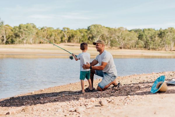 A young boy, dressed in shorts and t shirt, is facing the water while holding a fishing rod. A man kneels next to him, on the bank of the reservoir and holds the boy&rsquo;s back, smiling at him.